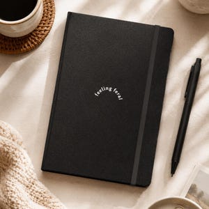 May include: A black notebook with a textured cover featuring the white text "feeling feral" in an arched design. The notebook includes a vertical black elastic closure band and is positioned next to a black ballpoint pen on a neutral surface.