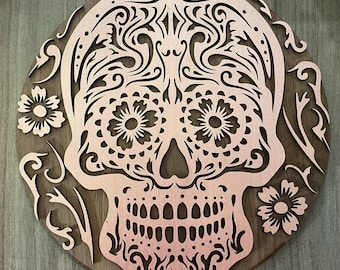 May include: A round wooden wall art piece featuring a laser-cut sugar skull design. The skull is light brown with intricate floral and swirl patterns, set against a darker brown background. Two flowers and decorative elements surround the skull.