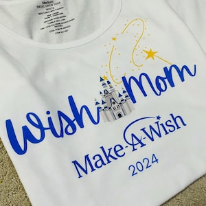 Pode incluir: Camiseta branca com gola redonda com o texto "Wish Mom Make-A-Wish 2024" em azul. Um castelo e estrelas amarelas tamb&eacute;m s&atilde;o apresentados na camiseta. A camiseta &eacute; de tamanho m&eacute;dio.