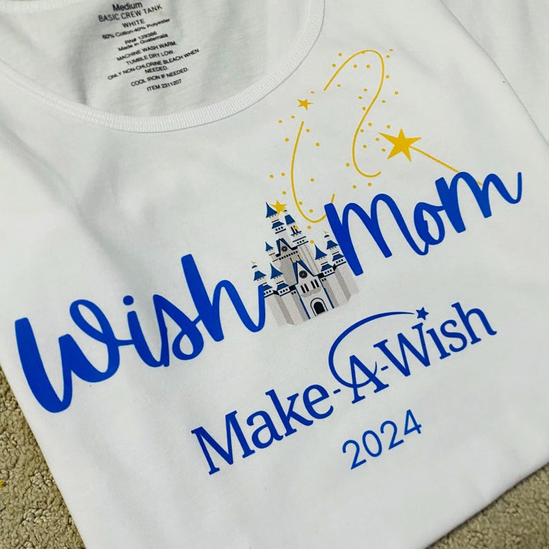 Pode incluir: Camiseta branca com gola redonda com o texto "Wish Mom Make-A-Wish 2024" em azul. Um castelo e estrelas amarelas tamb&eacute;m s&atilde;o apresentados na camiseta. A camiseta &eacute; de tamanho m&eacute;dio.