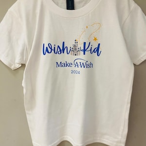 Pode incluir: Camiseta branca com as palavras "Wish Kid" em azul, um gr&aacute;fico de castelo e "Make-A-Wish 2024" impresso abaixo. Um desenho de estrelas douradas e amarelas est&aacute; acima das palavras. A camiseta est&aacute; pendurada em um cabide branco.