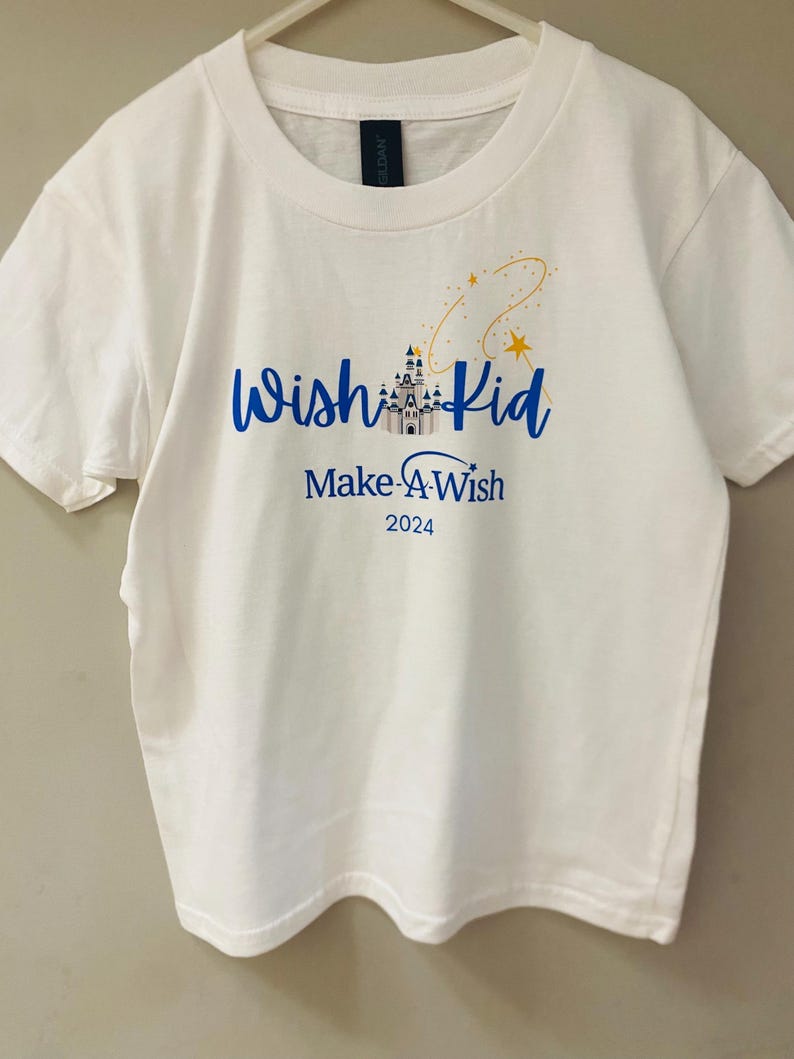 Pode incluir: Camiseta branca com as palavras "Wish Kid" em azul, um gr&aacute;fico de castelo e "Make-A-Wish 2024" impresso abaixo. Um desenho de estrelas douradas e amarelas est&aacute; acima das palavras. A camiseta est&aacute; pendurada em um cabide branco.