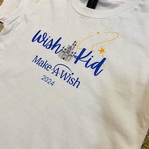 Pode incluir: Camiseta branca com texto azul que diz "wish kid" e "Make-A-Wish 2024". O design inclui uma ilustra&ccedil;&atilde;o de castelo e uma estrela amarela com uma linha curva.