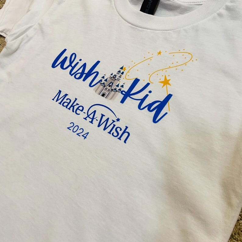 Pode incluir: Camiseta branca com texto azul que diz "wish kid" e "Make-A-Wish 2024". O design inclui uma ilustra&ccedil;&atilde;o de castelo e uma estrela amarela com uma linha curva.