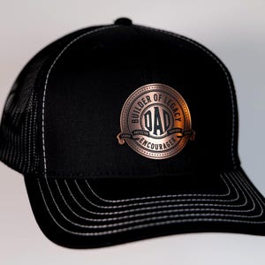May include: Black trucker hat with a copper-colored circular emblem. The emblem features the word "DAD" in the center, surrounded by the words "BUILDER OF LEGACY" and "ENCOURAGER". The hat has a mesh back and a curved brim with white stitching.