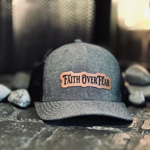 May include: A grey and black trucker hat with a brown leather patch that reads "Faith Over Fear".