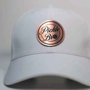 May include: A white baseball cap with a curved peak. The cap has a circular, copper-coloured patch with the words "Pickle Bro" in a script font, and a pickleball graphic. The cap is made of a smooth, solid material.