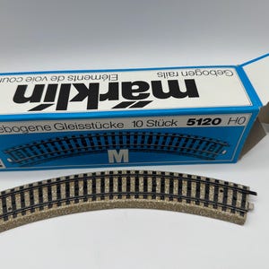Puede incluir: Una sección curva de vía de tren modelo con una base beige y rieles negros, junto a una caja azul y blanca. La caja dice "marklin" y "Gebogene Gleisstücke 10 Stück 5120 HO". La vía está diseñada para trenes modelo a escala HO.