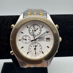 May include: A Seiko Quartz World Timer watch with a silver and gold-tone stainless steel strap. The white watch face features subdials and a date window. The bezel displays world cities.