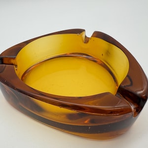 May include: A triangular amber glass ashtray with a smooth, rounded interior. The ashtray has a slightly raised rim and a deep well for holding cigarette ends.