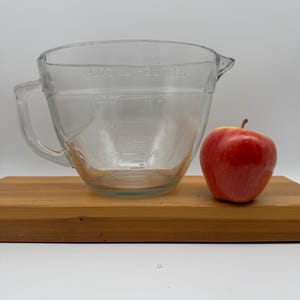 Op de afbeelding: Een heldere glazen maatbeker met een handvat en tuit, gemarkeerd met metingen tot 2000 ml of 2 liter. Een rode appel staat naast de beker op een houten snijplank. De beker is leeg.