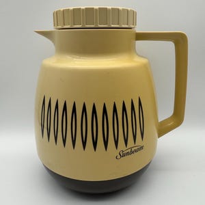 Op de afbeelding: Vintage Sunbeam thermoskan in crème en bruin. De thermoskan heeft een crèmekleurig deksel, handvat en body met een zwart ovaal patroon. De basis is bruin en het Sunbeam-logo is zichtbaar.