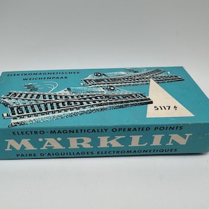 May include: A vintage Märklin box in teal features illustrations of model train tracks. The box reads "Electro-Magnetically Operated Points" and "5117 4". The box is for a pair of electro-magnetic points.