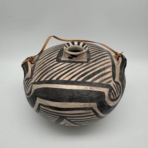 Puede incluir: Un recipiente de cerámica redondo hecho a mano con un patrón de rayas blancas y negras. El recipiente tiene una pequeña abertura en la parte superior y una correa de cuero para llevarlo. El diseño presenta formas y líneas geométricas.