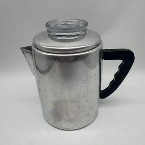 Puede incluir: Una cafetera de percolación de aluminio vintage con una tapa de vidrio transparente y un asa negra. La cafetera tiene un pico para verter y está diseñada para usar en la estufa. El metal tiene un acabado cepillado.