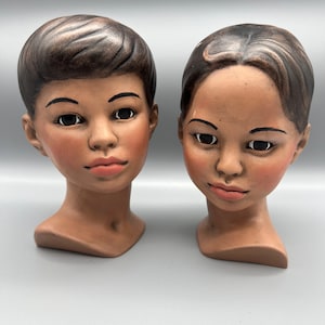 Könnte beinhalten: Zwei Keramikbüsten von Personen mit brauner Haut, schwarzen Haaren und bemalten Gesichtszügen. Die Büsten sind nach vorne gerichtet und haben einen neutralen Ausdruck.