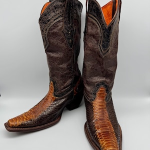 Puede incluir: Un par de botas de vaquero de cuero marrón con un estampado de piel de serpiente en el pie y una parte superior de cuero marrón oscuro. Las botas tienen una punta puntiaguda y un tacón apilado. El interior está forrado con tela naranja.