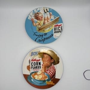 Op de afbeelding: Twee vintage Kellogg's ontbijtgranen borden. Eén bord toont een kind op een schommel met de tekst "Swing to Crispness" en "Kellogg's Rice Krispies". Het andere bord toont een jongen met een kom ontbijtgranen en de tekst "Kellogg's Corn Flakes."