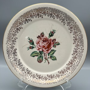 May include: A white ceramic dinner plate with a gold rim and a delicate gold floral pattern. The centre of the plate features a pink rose with green leaves.