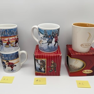May include: Three Tim Hortons ceramic mugs. Two mugs feature hockey scenes with players on ice. One mug is cream-coloured with a tan interior and a gold logo. Two mugs are stacked on the left, one sits on a red box, and one is inside a red box.