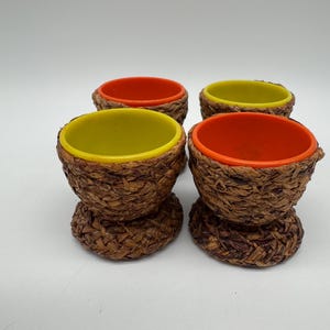 Könnte beinhalten: Vier dekorative Eierbecher. Jeder Becher hat eine gewebte, braune Außenseite und eine farbenfrohe Innenseite. Zwei haben orangefarbene Innenräume und zwei gelbe. Auf weißem Hintergrund.