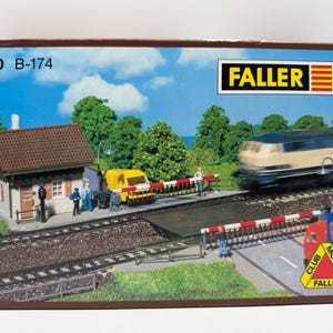 Op de afbeelding: Modeltreinsetdoos met een miniatuur spoorwegscène met een station, overweg en een snelle trein. De doos heeft de tekst "FALLER" en "HO B-174". De scène bevat een gele bestelwagen en miniatuurfiguren.