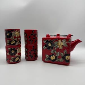 May include: A red ceramic tea set with a teapot and four cups. The set is decorated with black and gold floral designs. The teapot is square with a spout and handle. The cups are cylindrical.