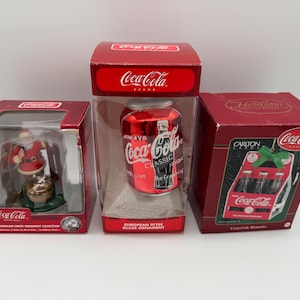 Può includere: Tre decorazioni natalizie a tema Coca-Cola in scatole. Uno presenta una figura di Babbo Natale, un altro una lattina rossa e il terzo un'esposizione di bottiglie in miniatura. Le scatole sono rosse con il logo Coca-Cola.