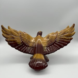 Könnte beinhalten: Eine Keramik-Adlerfigur mit brauner und beigefarbener Glasur, die Flügel im Flug ausgebreitet. Der Adler befindet sich in einer Flugposition mit dem Kopf zur Seite gedreht.