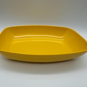 Puede incluir: Una fuente de servir rectangular de color amarillo brillante con esquinas redondeadas. La fuente tiene un acabado liso y brillante y una forma ligeramente curvada. La fuente vacía está sobre una superficie blanca.