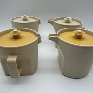 Op de afbeelding: Vier crèmekleurige vintage kannen met mosterdgele deksels en handvatten. Elke kan heeft een tuit en een kleine knop op het deksel. De kannen zijn in een groep gerangschikt, waarbij hun ontwerp wordt getoond.