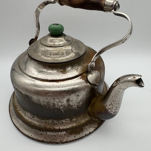 May include: A vintage silver-plated tea kettle with a wooden handle and a green knob on the lid. The kettle has a worn, aged appearance.