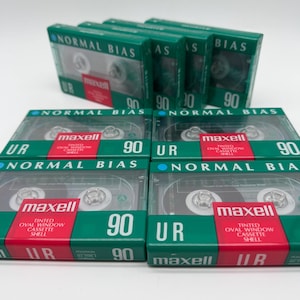 Puede incluir: Una colección de cintas de casete de audio Maxell UR 90. Cada cinta está en una caja verde y roja con el texto "NORMAL BIAS" y "90" impreso. La cinta de casete es visible a través de una ventana transparente.