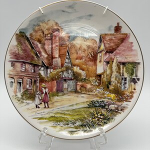 Puede incluir: Un plato de cerámica blanco con un borde dorado presenta una pintura de acuarela de una pintoresca escena de un pueblo inglés. La pintura representa una calle bordeada de casas de ladrillo, vegetación exuberante y dos figuras caminando por el sendero.