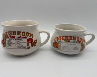 Puede incluir: Dos tazas de sopa de cerámica blanquecina. Una taza tiene "MUSHROOM" en letras marrones con una receta. La otra taza tiene "CHICKEN SOUP" en letras naranjas con una receta. Ambas tazas tienen asas y están decoradas con ilustraciones.