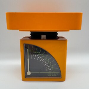 May include: An orange kitchen scale with a black dial and white markings. The scale has a rectangular base and a tray on top. The dial shows measurements in ounces and grams. The brand name is visible on the bottom of the dial.