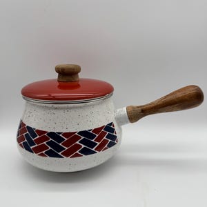 May include: A white speckled saucepan with a red lid and wooden handle. The pot features a woven pattern in red and navy blue. The lid has a wooden knob. The handle is made of wood.