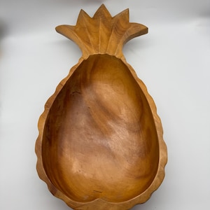 May include: A wooden pineapple-shaped bowl with a natural wood finish. The bowl has a deep interior and a detailed, carved exterior, including the pineapple crown. The bowl is designed for serving or display.