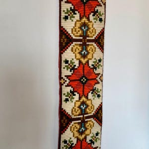 May include: A long, rectangular tapestry with a repeating pattern of red, white, brown, and yellow flowers. The tapestry is hanging on a white wall with a black metal hook.