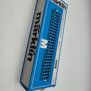 May include: A blue and white Märklin box containing straight track sections for model trains. The box features the Märklin logo and product information, including "5106" and "HO". The box is angled, showing the side with the text "Straight track sections".