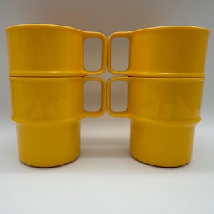 Puede incluir: Cuatro tazas de color amarillo brillante apiladas en pares. Cada taza tiene un cuerpo redondeado, un asa y un borde ligeramente ensanchado. Las tazas están hechas de un material brillante y están sobre un fondo blanco liso.