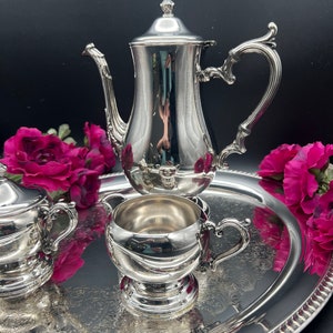 Op de afbeelding: Een zilveren theeservies met een theepot, roomkan en suikerpot op een zilveren dienblad. De set is versierd met een sierlijk bloemenpatroon.