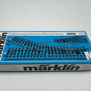 May include: A blue Märklin box featuring a diagram of model train tracks and the text "Elektromagnetisches Weichenpaar 5202 HO". The box also includes the text "Pair of solenoid-operated turnouts" and "Made in Western Germany".