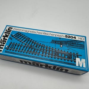 May include: A blue Märklin box featuring a diagram of model train tracks. The box has the text "Elektromagnetisches Weichenpaa2x5204 HO" and the Märklin logo. The box is for model train enthusiasts.