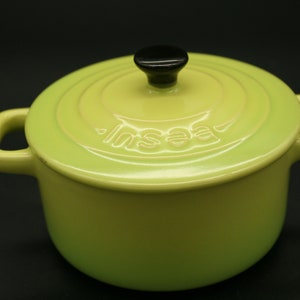 以下が含まれることがあります： ふたに黒いノブが付いた緑色のセラミック製の鍋。鍋には持ち手が2つあり、ふたには「Insa」という文字が印刷されています。