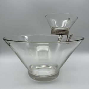 Pode incluir: Um conjunto de servir de vidro transparente para batatas fritas e molhos. A tigela maior tem uma forma cónica larga, e a tigela menor é suspensa acima com um suporte de metal prateado. O conjunto foi concebido para servir snacks.