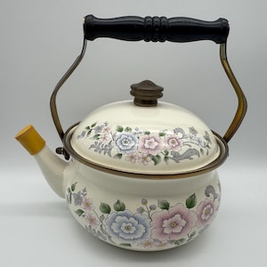 May include: Cream-colored enamel tea kettle with floral design in pink, blue, and green. The kettle has a black wooden handle, a brass-colored frame, and a yellow spout. A decorative kitchenware item.