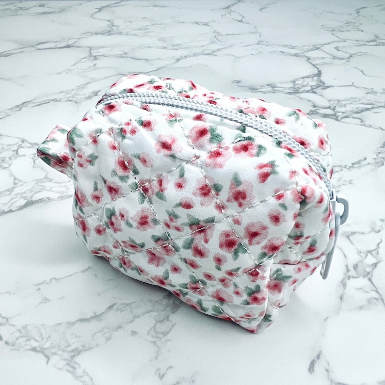 K&ouml;nnte beinhalten: Eine wei&szlig;e gesteppte Kosmetiktasche mit Rei&szlig;verschluss und einem Blumendruck mit rosa Bl&uuml;ten auf wei&szlig;em Hintergrund.