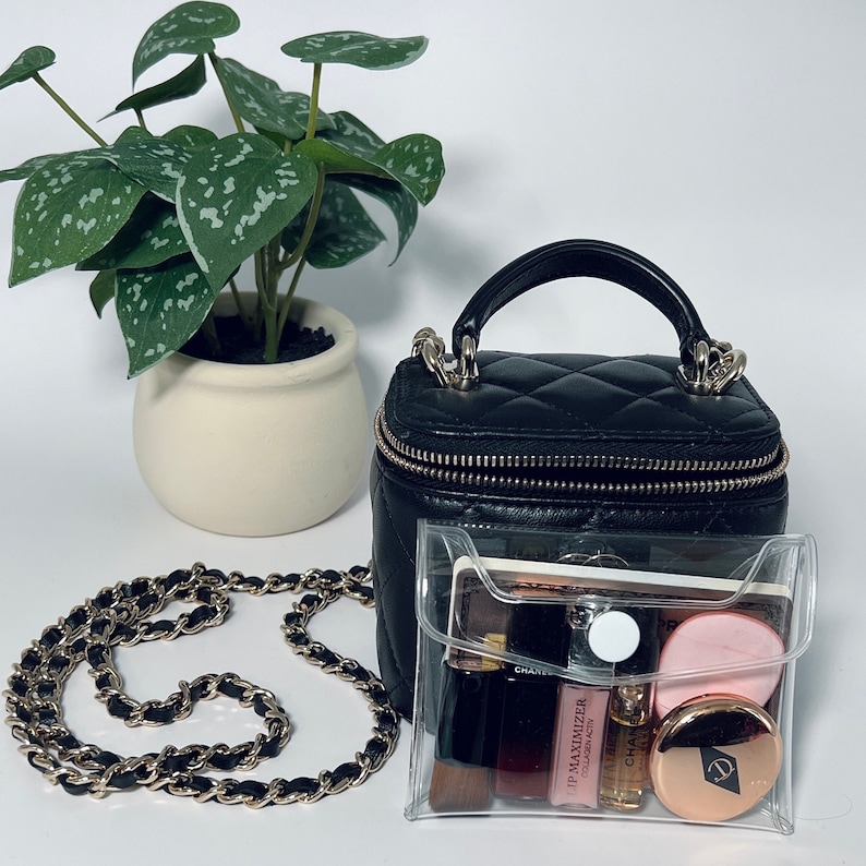 Puede incluir: Un bolso de mano de cuero negro acolchado con asa superior y correa de cadena dorada. Una bolsa de cosm&eacute;ticos transparente contiene art&iacute;culos de maquillaje. Una peque&ntilde;a planta en maceta est&aacute; en el fondo.