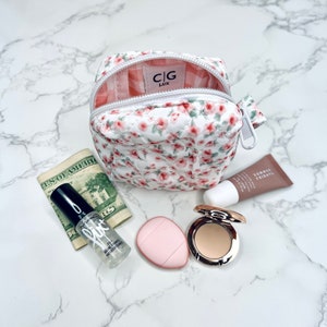 K&ouml;nnte beinhalten: Eine kleine wei&szlig;e, gesteppte Kosmetiktasche mit Blumenmuster, Rei&szlig;verschluss und rosa-wei&szlig;em Karomuster. Die Tasche ist offen und enth&auml;lt eine durchsichtige Flasche mit Make-up-Fixierspray, einen rosa Make-up-Schwamm, eine kompakte Foundation und eine Tube Lippenbalsam.